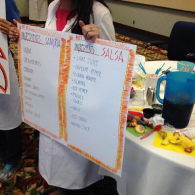A team member holds up ingredient lists for Sangria and Salsa at the Salsa Showdown team building event, with colorful ingredients and materials on the table. thumbnail