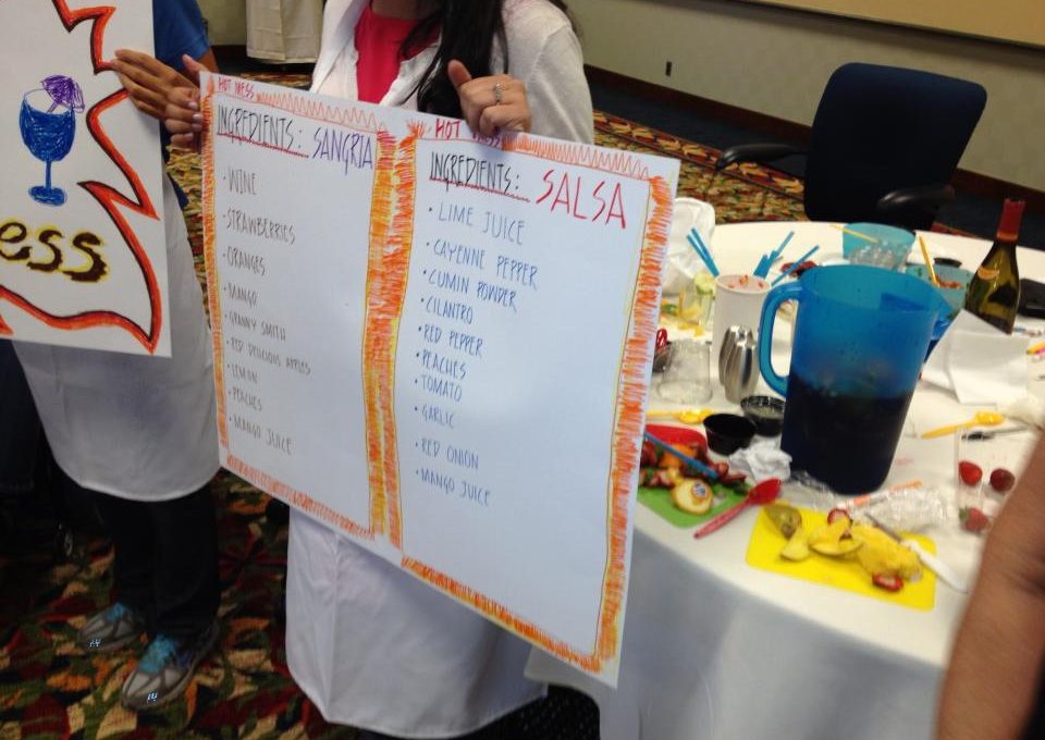 A team member holds up ingredient lists for Sangria and Salsa at the Salsa Showdown team building event, with colorful ingredients and materials on the table.