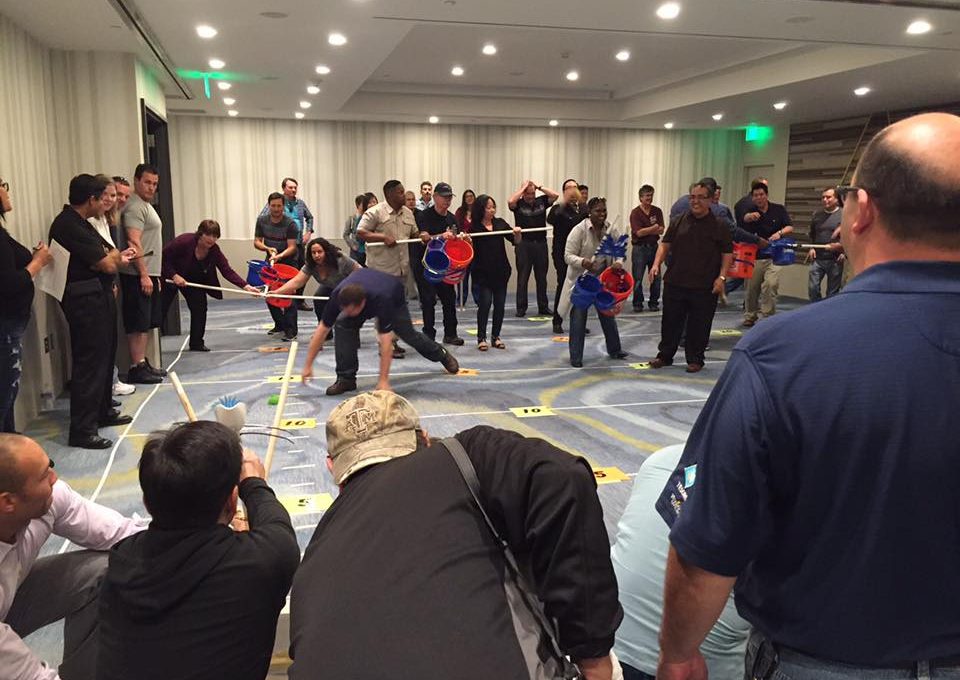 Participants in an indoor setting engaging in a competitive team building activity with catapults, buckets, and various objects, demonstrating teamwork, strategy, and coordination during the Catapult to Success event.