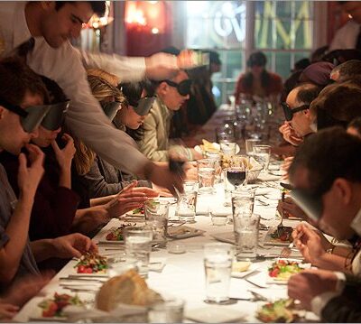 Large group of participants at a 'Dining in the Dark' event seated around a long dining table, wearing blindfolds as they enjoy a meal served by waitstaff, focusing on enhancing their sensory experience through taste and smell. thumbnail