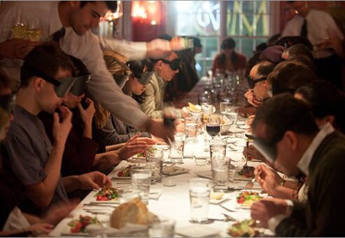 Large group of participants at a 'Dining in the Dark' event seated around a long dining table, wearing blindfolds as they enjoy a meal served by waitstaff, focusing on enhancing their sensory experience through taste and smell.