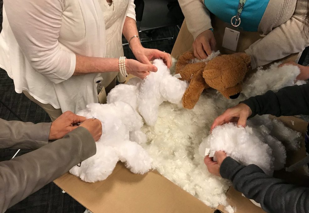 A group of participants are seen from above working together to stuff teddy bears. Their hands are holding various plush bear parts while reaching into a large cardboard box filled with stuffing material. The teddy bears are in different stages of assembly, with some already partially stuffed. The focus is on teamwork and collaboration as they fill the bears for the Team Teddy Rescue Bear team building event.