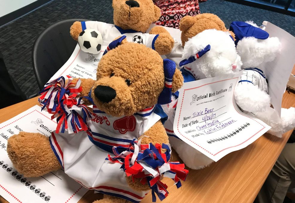 A collection of completed teddy bears dressed in sports-themed outfits, including one as a cheerleader with pom-poms and another as a soccer player with a soccer ball. Each bear has an 'Official Birth Certificate' placed underneath it with handwritten details like the bear's name and date of birth. The scene captures the results of a teddy bear-building activity, part of a charitable team building event.