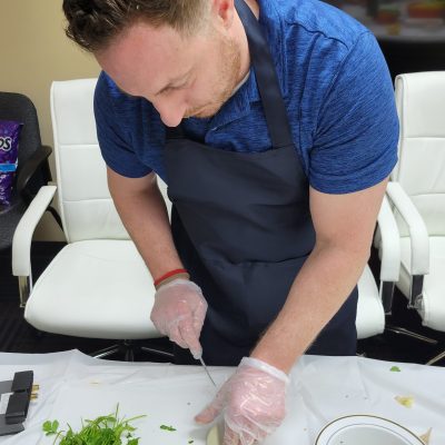 A man in a blue shirt and black apron is carefully chopping an onion at a table during a team building chili cook-off event. He is wearing disposable gloves, and there are herbs, chopped vegetables, and a small bowl with ingredients on the table in front of him. thumbnail