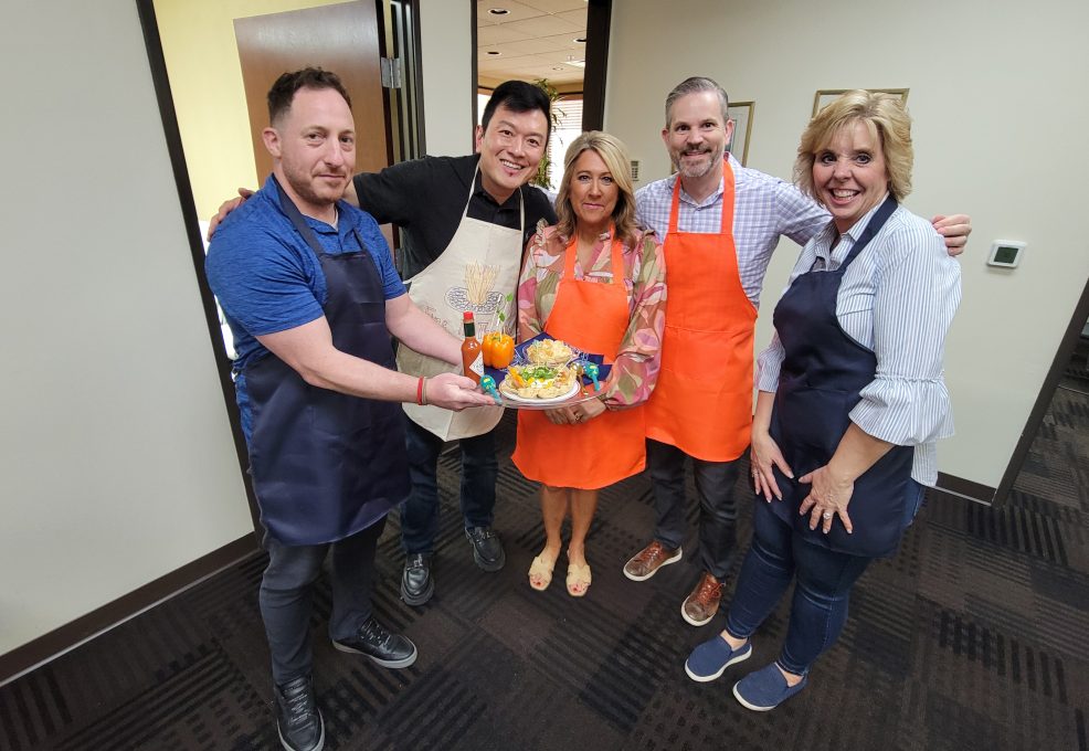 A group of five people standing together, smiling and holding a dish they prepared during a team building chili cook-off event. The group is standing indoors, with each person wearing an apron of different colors (blue, orange, and white). They are posing proudly with their food creation, likely showcasing it for the competition. The atmosphere is lighthearted, and the participants appear happy and engaged.