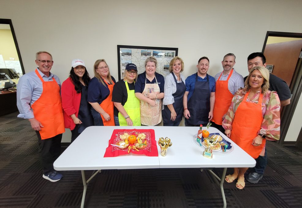 A group of individuals stands together, smiling, behind a table at a team building chili cook-off event. Some are wearing brightly colored aprons in shades of orange, blue, and green. The table in front of them displays two creatively presented chili dishes, along with small golden trophies.