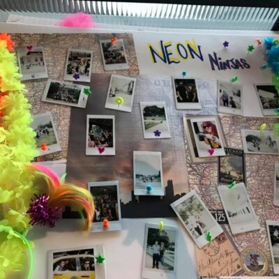 A vibrant display board titled 'Neon Ninjas' from a Polaroid scavenger hunt, decorated with colorful leis and neon accents. The board features a collection of Polaroid photos pinned around a central map, capturing fun moments from the scavenger hunt challenges. The lively decorations and creative photo arrangement highlight the energy and excitement of the team building event. thumbnail