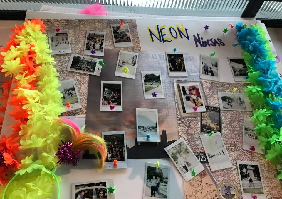 A vibrant display board titled 'Neon Ninjas' from a Polaroid scavenger hunt, decorated with colorful leis and neon accents. The board features a collection of Polaroid photos pinned around a central map, capturing fun moments from the scavenger hunt challenges. The lively decorations and creative photo arrangement highlight the energy and excitement of the team building event.