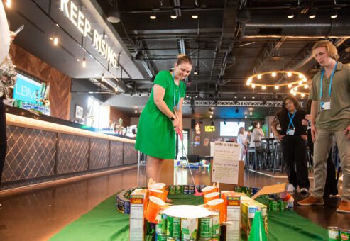 A participant plays on a creatively designed mini golf course made from canned goods and food packages during a 'Build Your Own Mini Golf Course' team building event. The event takes place in a lively venue, combining fun, teamwork, and charity, with the food items used for the course being donated to a local food pantry afterward.