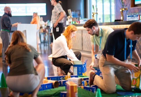 Participants collaborate to build a mini golf course using canned goods and packaged food items during a 'Build Your Own Mini Golf Course' team building event. The activity encourages teamwork and creativity, with the food items being donated to a local food pantry after the event, combining fun with a charitable cause.