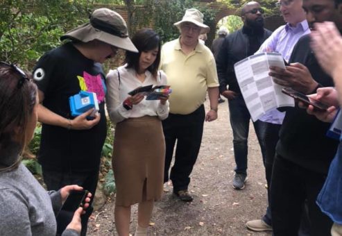 A group of team members gathered in a garden, reviewing clues and maps during an outdoor scavenger hunt. Each person is focused on their materials, including instant cameras and printed instructions, as they collaborate to solve challenges.
