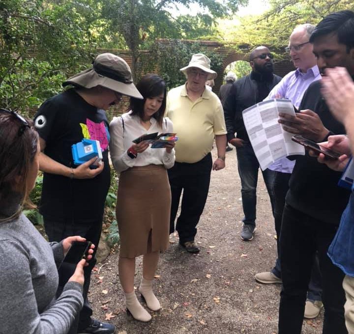 A group of team members gathered in a garden, reviewing clues and maps during an outdoor scavenger hunt. Each person is focused on their materials, including instant cameras and printed instructions, as they collaborate to solve challenges.