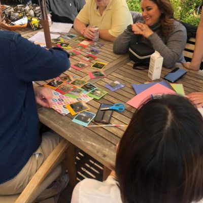 A group of team members gathered around a wooden table outdoors, collaborating on a photo collage as part of a Polaroid scavenger hunt. The table is filled with colorful paper, Polaroid photos, scissors, and crafting materials as participants engage in friendly conversation and creative teamwork, enjoying a relaxing and productive moment in a sunny garden setting. thumbnail