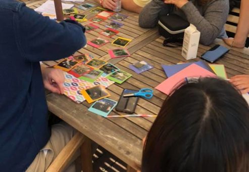 A group of team members gathered around a wooden table outdoors, collaborating on a photo collage as part of a Polaroid scavenger hunt. The table is filled with colorful paper, Polaroid photos, scissors, and crafting materials as participants engage in friendly conversation and creative teamwork, enjoying a relaxing and productive moment in a sunny garden setting.