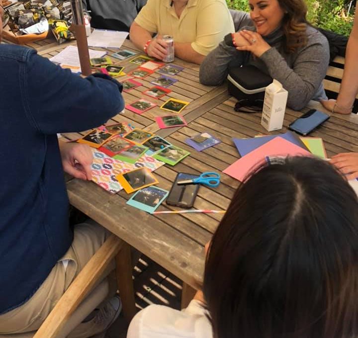 A group of team members gathered around a wooden table outdoors, collaborating on a photo collage as part of a Polaroid scavenger hunt. The table is filled with colorful paper, Polaroid photos, scissors, and crafting materials as participants engage in friendly conversation and creative teamwork, enjoying a relaxing and productive moment in a sunny garden setting.