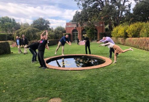 A group of team members engage in a playful pose around a small circular pond in a lush garden during an outdoor team building event.