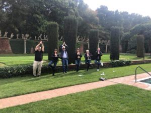 A group of team members standing in a landscaped garden, striking synchronized playful poses with hands above their heads during an outdoor team building event. They stand in front of neatly trimmed hedges and greenery, creating a lighthearted and engaging moment in a scenic, natural environment.