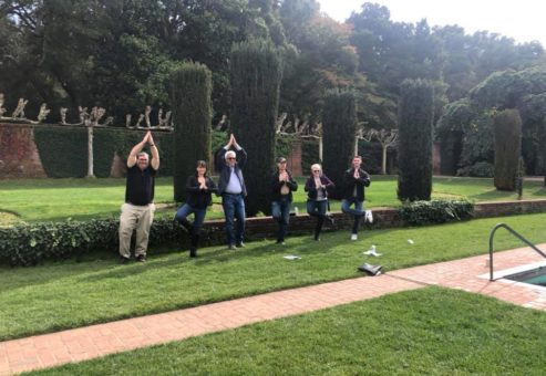 A group of team members standing in a landscaped garden, striking synchronized playful poses with hands above their heads during an outdoor team building event. They stand in front of neatly trimmed hedges and greenery, creating a lighthearted and engaging moment in a scenic, natural environment.