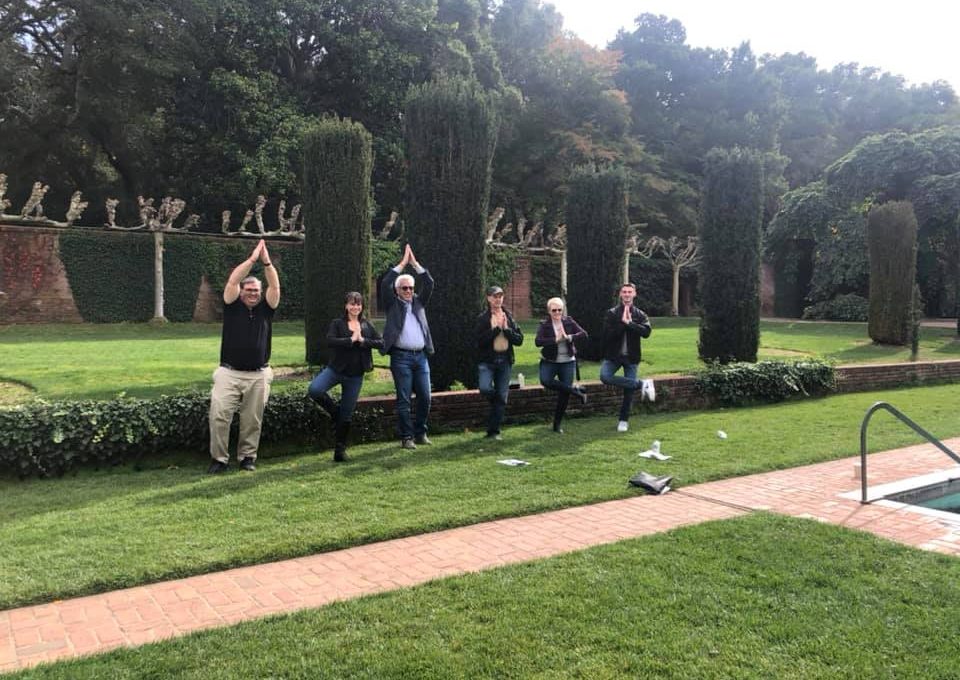 A group of team members standing in a landscaped garden, striking synchronized playful poses with hands above their heads during an outdoor team building event. They stand in front of neatly trimmed hedges and greenery, creating a lighthearted and engaging moment in a scenic, natural environment.