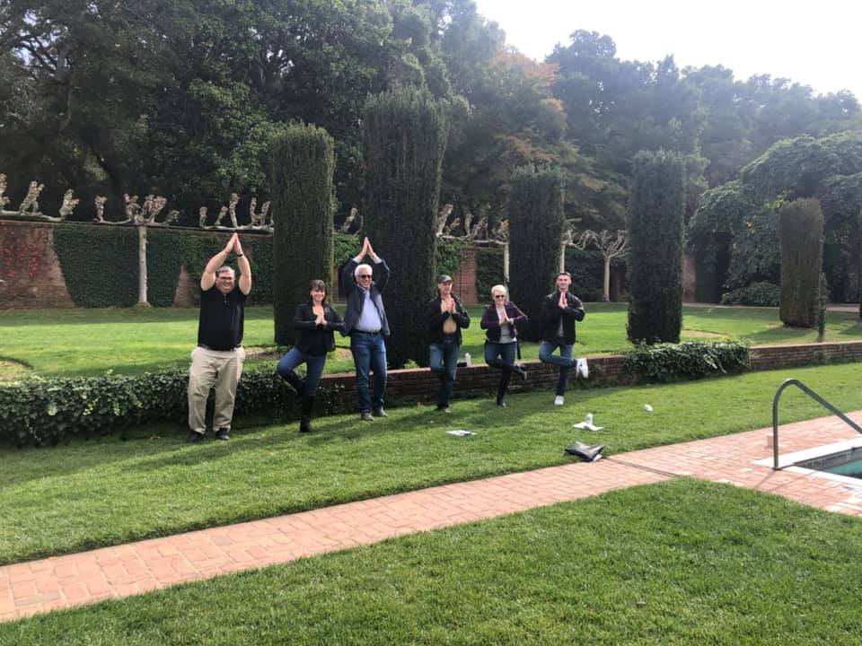 A group of team members standing in a landscaped garden, striking synchronized playful poses with hands above their heads during an outdoor team building event. They stand in front of neatly trimmed hedges and greenery, creating a lighthearted and engaging moment in a scenic, natural environment.