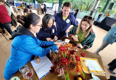 Four team members engage in a fun Polaroid scavenger hunt, reviewing instant photos taken during an outdoor team building event. The group gathers around a table with vibrant fall-themed flower arrangements and drinks.