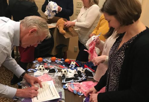 A group of participants actively engage in assembling teddy bears during the Team Teddy Rescue Bear team building event. They are working around a table filled with teddy bear parts, small outfits, and soccer ball accessories. One person is filling out a certificate, while others are selecting outfits and assembling the bears.