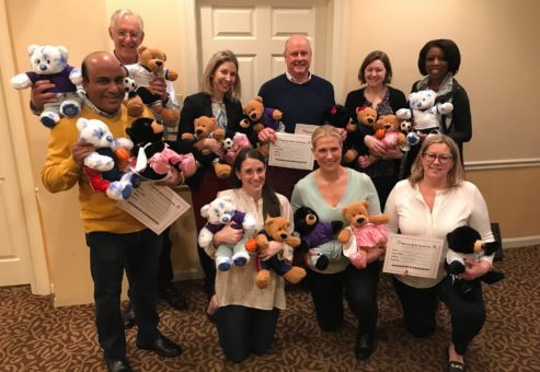 A group of nine smiling participants proudly display the teddy bears they have created during the Team Teddy Rescue Bear team building event. Each person holds one or more stuffed bears in various outfits and colors.
