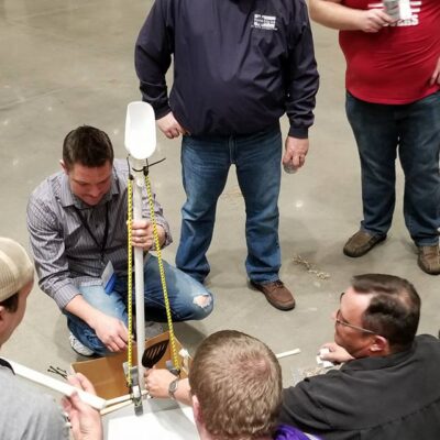 A group of participants indoors gathered around a table, actively assembling a catapult as part of the Catapult to Success team building activity, focusing on collaboration, innovation, and hands-on problem-solving. thumbnail