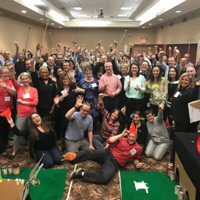 A large group of participants celebrating together after a successful 'Build Your Own Mini Golf Course' team building event. The group poses with their hands raised and mini golf clubs in hand, surrounded by creative mini golf holes made from canned goods and other non-perishable food items. thumbnail