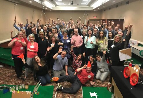 A large group of participants celebrating together after a successful 'Build Your Own Mini Golf Course' team building event. The group poses with their hands raised and mini golf clubs in hand, surrounded by creative mini golf holes made from canned goods and other non-perishable food items.