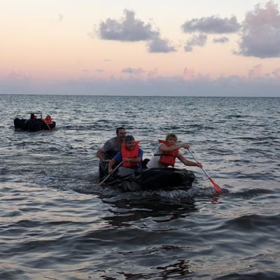 Teams paddling cardboard boats on the open water during the Build-A-Boat team building challenge at sunset. thumbnail