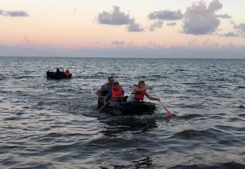 Teams paddling cardboard boats on the open water during the Build-A-Boat team building challenge at sunset.