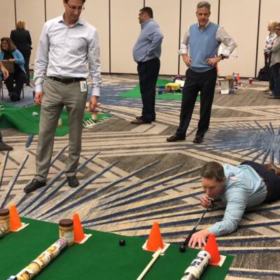 A participant lying on the floor carefully lines up a shot during a 'Build Your Own Mini Golf Course' team building event. The mini golf course is constructed from canned goods and other food items, promoting both fun teamwork and charity, with the food being donated to a local pantry afterward. thumbnail