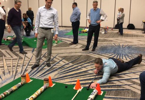 A participant lying on the floor carefully lines up a shot during a 'Build Your Own Mini Golf Course' team building event. The mini golf course is constructed from canned goods and other food items, promoting both fun teamwork and charity, with the food being donated to a local pantry afterward.