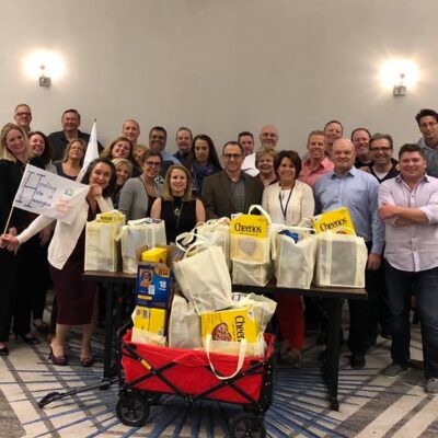A group of participants proudly stands behind a collection of bags filled with groceries after a successful 'Build Your Own Mini Golf Course' team building event. The event encourages teamwork and charitable giving, with the food items being donated to a local food pantry to support families in need. thumbnail