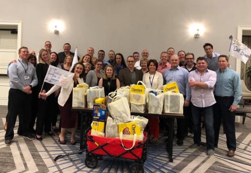A group of participants proudly stands behind a collection of bags filled with groceries after a successful 'Build Your Own Mini Golf Course' team building event. The event encourages teamwork and charitable giving, with the food items being donated to a local food pantry to support families in need.