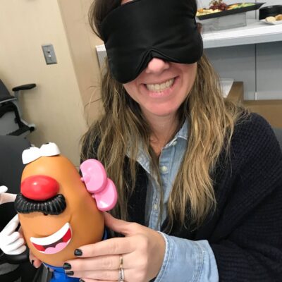 A woman smiling and holding a Mr. Potato Head toy while wearing a black blindfold. She is participating in a fun activity as part of the Toys for Tykes team building event, where participants assemble a toy blindfolded as a challenge. thumbnail