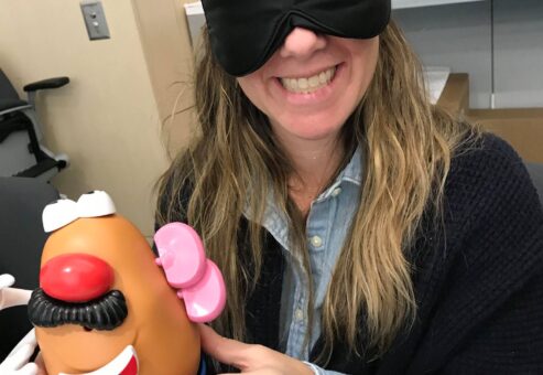 A woman smiling and holding a Mr. Potato Head toy while wearing a black blindfold. She is participating in a fun activity as part of the Toys for Tykes team building event, where participants assemble a toy blindfolded as a challenge.