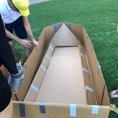 Team members constructing a cardboard boat framework during the Build-A-Boat team building challenge on a grassy outdoor space. thumbnail