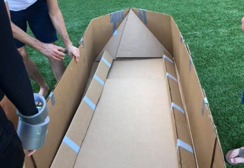 Team members constructing a cardboard boat framework during the Build-A-Boat team building challenge on a grassy outdoor space.