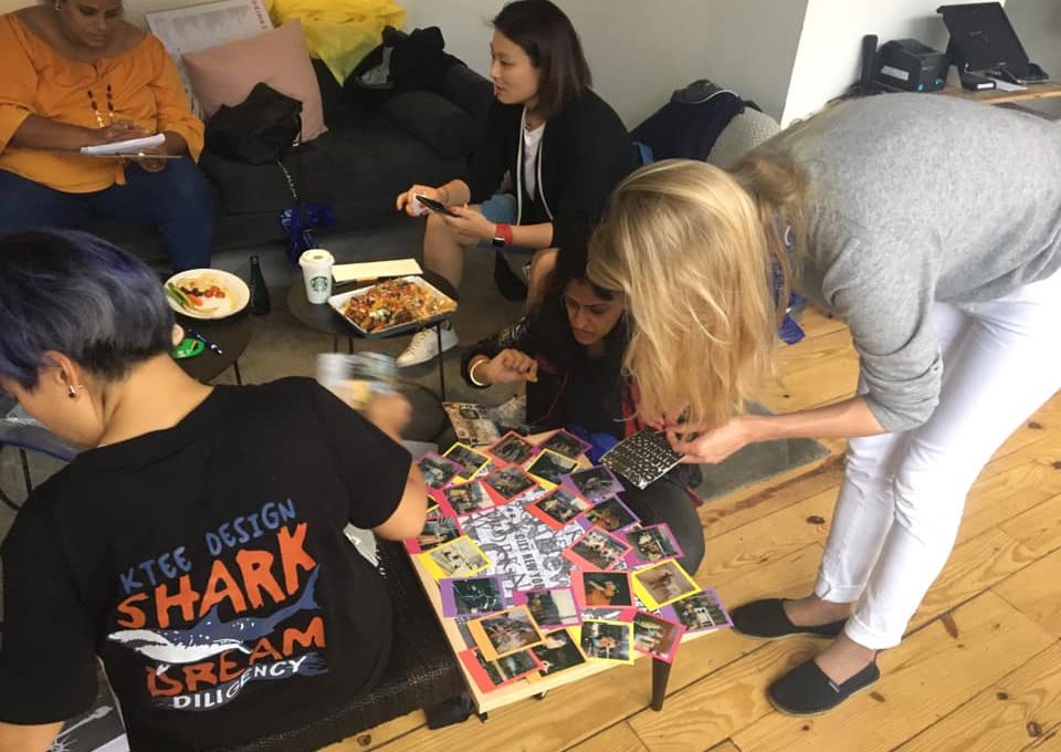 A dynamic scene of team members assembling a collage on the floor, using Polaroid photos from a team building scavenger hunt. Individuals focus on arranging the photos, and enjoying light snacks. The activity fosters teamwork and creativity.