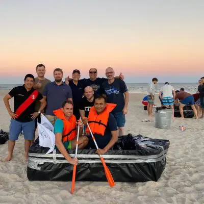 Team poses with their completed cardboard boat on the beach during the Build-A-Boat team building event at sunset. thumbnail