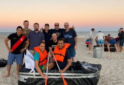 Team poses with their completed cardboard boat on the beach during the Build-A-Boat team building event at sunset.