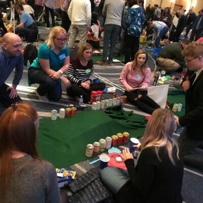 A group of participants collaborating on building a mini golf course using canned goods and non-perishable food items during a 'Build Your Own Mini Golf Course' team building event. thumbnail