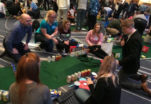 A group of participants collaborating on building a mini golf course using canned goods and non-perishable food items during a 'Build Your Own Mini Golf Course' team building event.