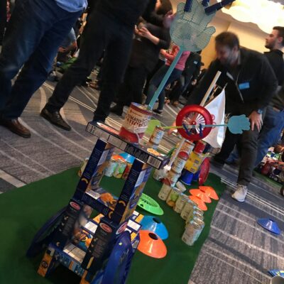 A creatively designed mini golf hole built from food cans, pasta boxes, and other non-perishable items during a 'Build Your Own Mini Golf Course' team building event. The course features a playful structure with a toy wheel and hand decorations. thumbnail