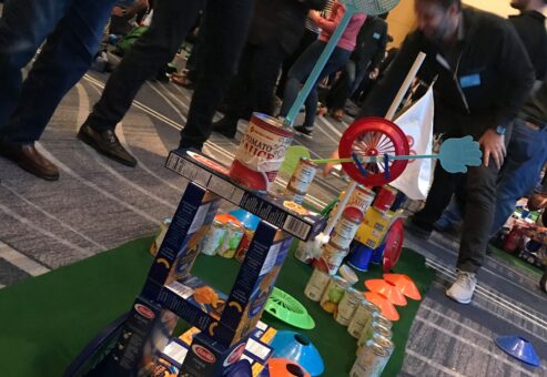 A creatively designed mini golf hole built from food cans, pasta boxes, and other non-perishable items during a 'Build Your Own Mini Golf Course' team building event. The course features a playful structure with a toy wheel and hand decorations.
