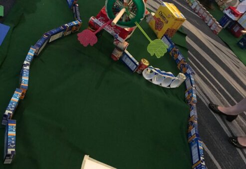 A creative mini golf course built from food packages, including cereal boxes, cans, and snack items, with a 'Start' sign marking the beginning of the hole. Participants designed the course as part of a 'Build Your Own Mini Golf Course' team building event, combining fun, creativity, and charity. The food items used are later donated to a local pantry.