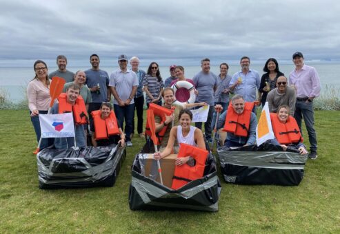 Team posing with their decorated cardboard boats and flags during the Build-A-Boat team building challenge near the water.
