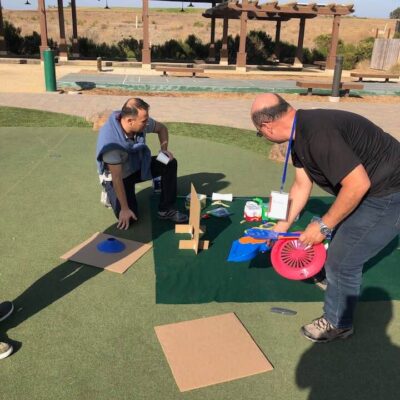 Two participants engaged in a team building 'Build Your Own Mini Golf Course' event, working on constructing a mini golf hole using cardboard, a fan, and other creative materials on a mat laid out on a grassy area outdoors. This activity encourages teamwork and innovation while using everyday objects to create a golf course.. thumbnail